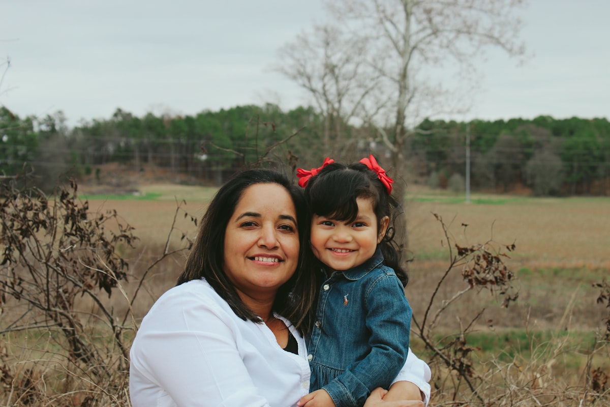 A mother and her young daughter smiling together in an open field.